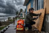 Una mujer con linterna frontal clavando madera contrachapada en una casa costera bajo un cielo tormentoso amenazante. A su lado, una mochila de emergencia naranja completamente equipada con botellas de agua, botiquín de primeros auxilios y suministros.