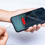 Manos sosteniendo un celular con un icono rojo de batería baja en pantalla y un cable de carga blanco junto al dispositivo.