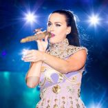 Katy Perry canta con micrófono dorado y vestido lila con adornos brillantes durante un concierto iluminado con luces azules.