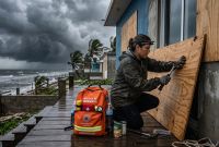 Una mujer con linterna frontal clavando madera contrachapada en una casa costera bajo un cielo tormentoso amenazante. A su lado, una mochila de emergencia naranja completamente equipada con botellas de agua, botiquín de primeros auxilios y suministros.