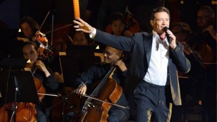 Hugh Jackman canta con micrófono y traje oscuro sobre un escenario, acompañado por músicos de una orquesta.