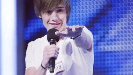 Liam Payne adolescente con camiseta blanca y micrófono en la mano durante su audición en The X Factor, frente a un fondo azul.