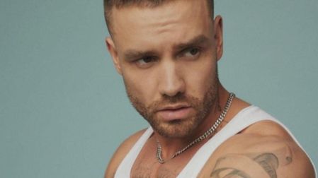 Liam Payne con camiseta blanca sin mangas, cadena plateada, barba corta y tatuajes en el hombro, frente a un fondo azul claro.