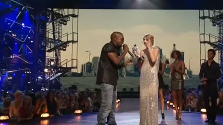 Kanye West con micrófono frente a Taylor Swift, vestida de plateado y sosteniendo su premio MTV en el escenario.