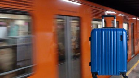 Maleta azul frente a un tren naranja del Metro de CDMX en movimiento dentro de una estación.