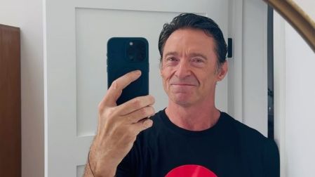 Hugh Jackman con camiseta negra y círculo rojo, sonríe mientras se toma una selfie frente a un espejo.