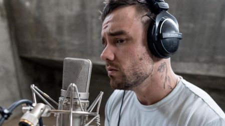 Liam Payne con audífonos negros y camiseta gris canta frente a un micrófono de estudio, con tatuajes visibles en el cuello.