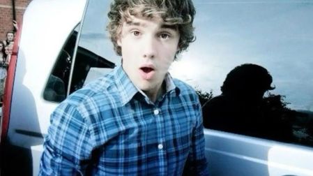Liam Payne de adolescente, con cabello ondulado y camisa azul a cuadros, junto a una camioneta blanca.