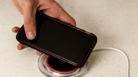 Mano colocando un celular negro con pantalla apagada sobre una base circular de carga inalámbrica conectada a un cable blanco