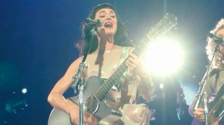 Katy Perry canta y toca una guitarra acústica durante un concierto, con luces intensas y otra música al fondo.