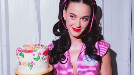 Katy Perry luce blusa rosa a cuadros y coletas con lazos rosas mientras sostiene un pastel decorado con flores y velas.