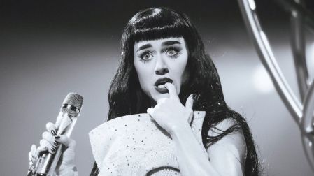 Katy Perry sostiene un micrófono y luce cabello negro con flequillo y vestido brillante durante una presentación en blanco y negro.