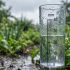 Una vista de primer plano de un pluviómetro de plástico transparente montado en un poste de madera en un jardín exuberante, con agua de lluvia acumulada midiendo aproximadamente 75 milímetros en la escala.