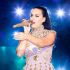 Katy Perry canta con micrófono dorado y vestido lila con adornos brillantes durante un concierto iluminado con luces azules.