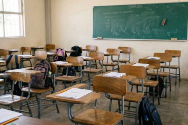 Vista interior de un aula de escuela vacía con filas de pupitres de madera, mochilas dejadas en los asientos y una pizarra verde al fondo.