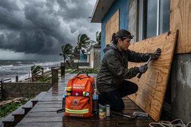 Una mujer con linterna frontal clavando madera contrachapada en una casa costera bajo un cielo tormentoso amenazante. A su lado, una mochila de emergencia naranja completamente equipada con botellas de agua, botiquín de primeros auxilios y suministros.