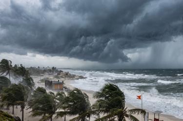 Fuerte oleaje y vientos intensos golpeando una zona costera durante el acercamiento de una tormenta tropical, con cielo oscuro y palmeras doblándose por las ráfagas.