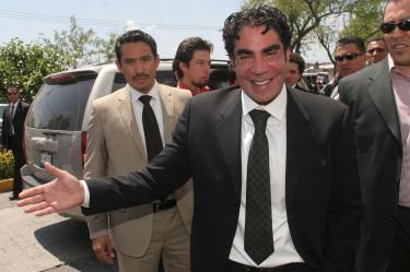 Jorge Kahwagi sonriendo de traje negro durante un evento político, rodeado de colaboradores y equipo de seguridad.