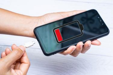Manos sosteniendo un celular con un icono rojo de batería baja en pantalla y un cable de carga blanco junto al dispositivo.