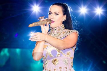 Katy Perry canta con micrófono dorado y vestido lila con adornos brillantes durante un concierto iluminado con luces azules.